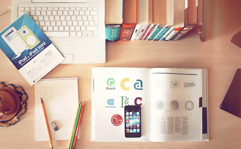 A desk laid out with a laptop, magazines, a notebook, and pencils